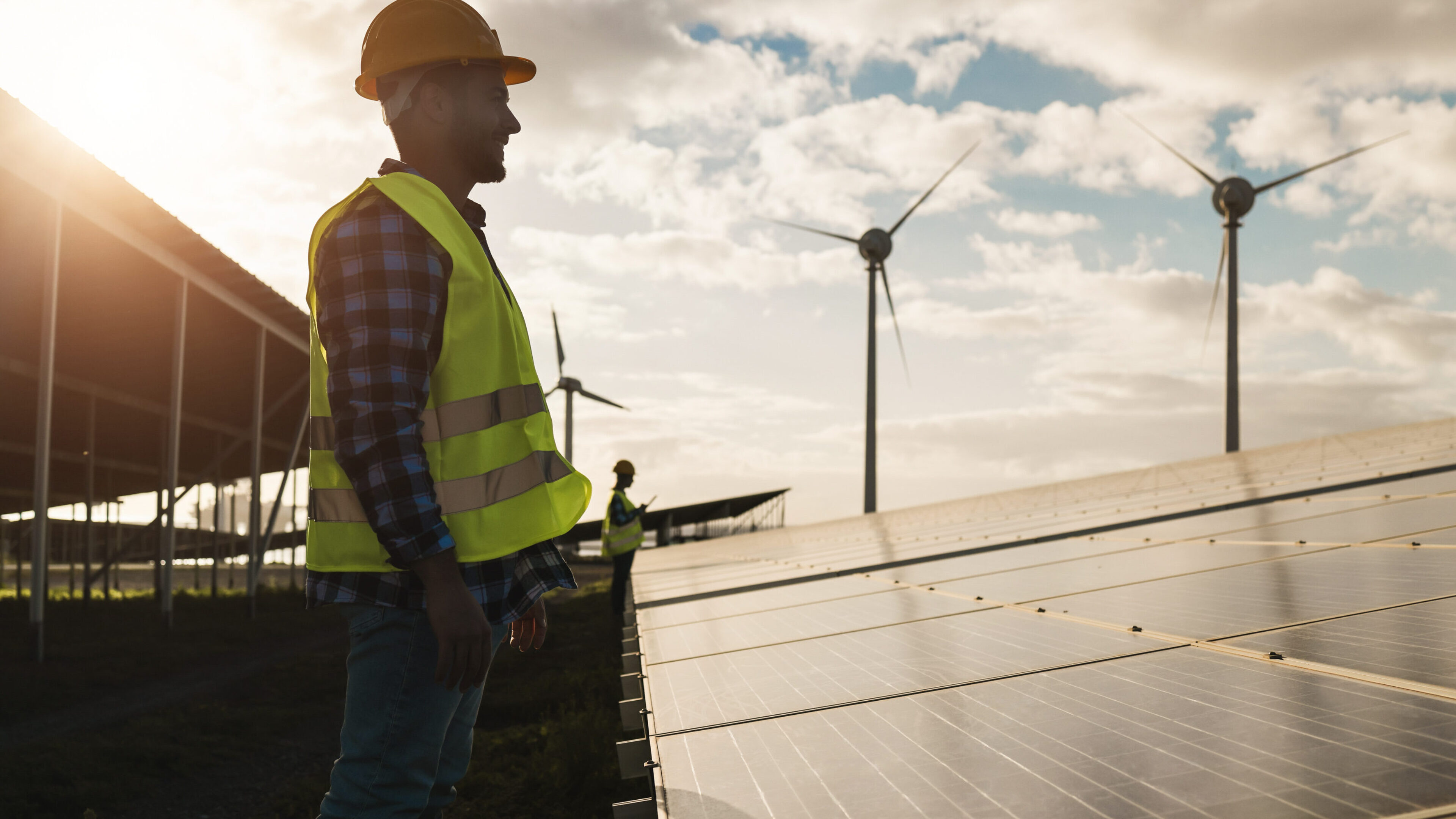 Froschperspektive eines Mannes in Sicherheitskleidung, der gerade an Solarparneelen und Windenergieanlagen arbeitet