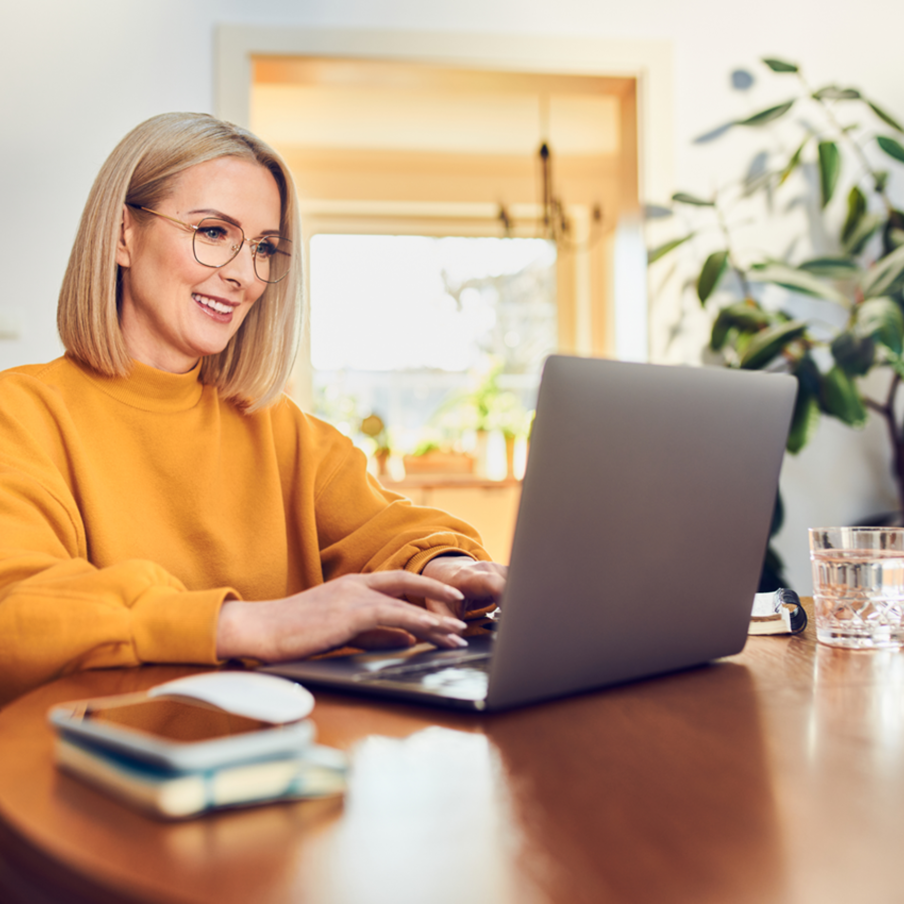 Eine blonde Frau mit Brille und gelbem Pullover sitzt an einem runden Tisch und arbeitet an ihrem Laptop.