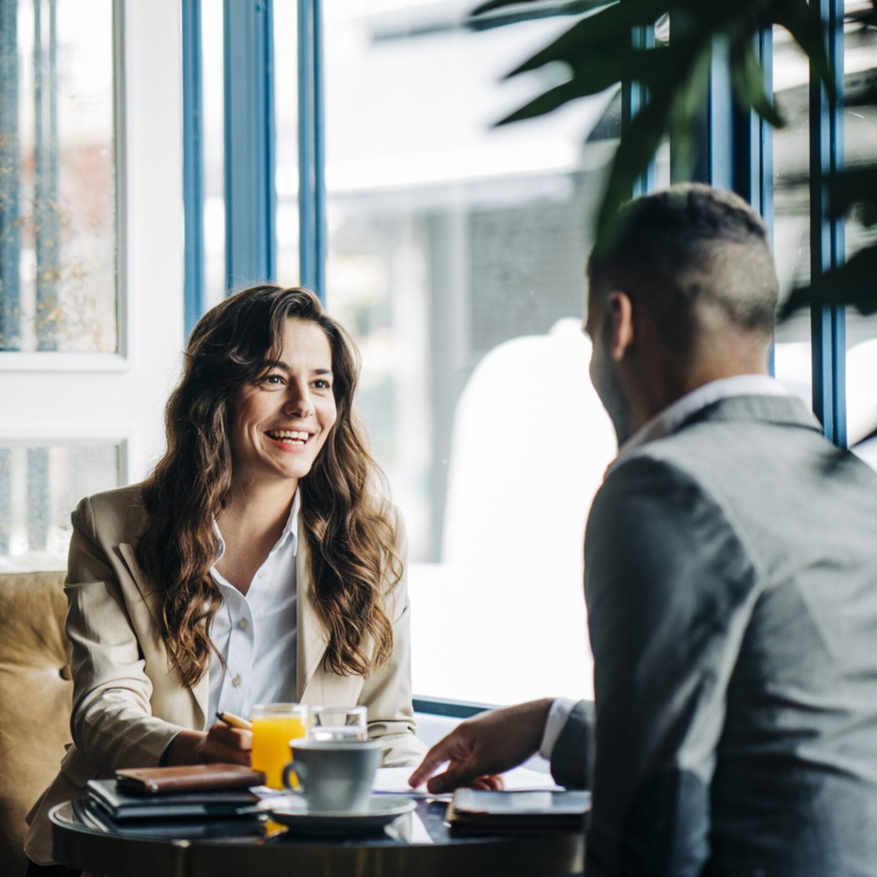 Zwei Personen sitzen in einem Café an einem kleinen Tisch und führen ein Gespräch, während Getränke und Unterlagen vor ihnen liegen.