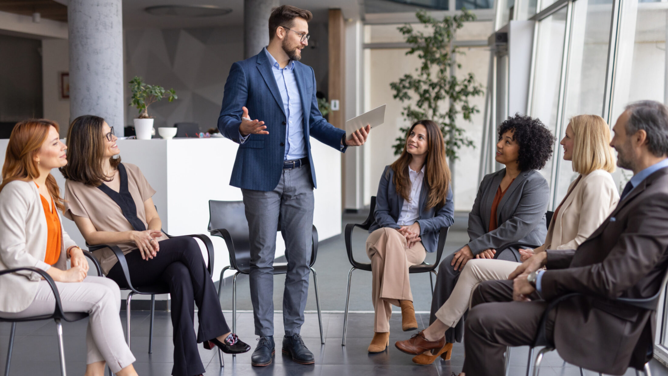 Eine Gruppe von Personen sitzt in einem Stuhlkreis, während eine stehende Person die Runde anleitet und ein Führungsgespräch oder Team-Meeting moderiert.