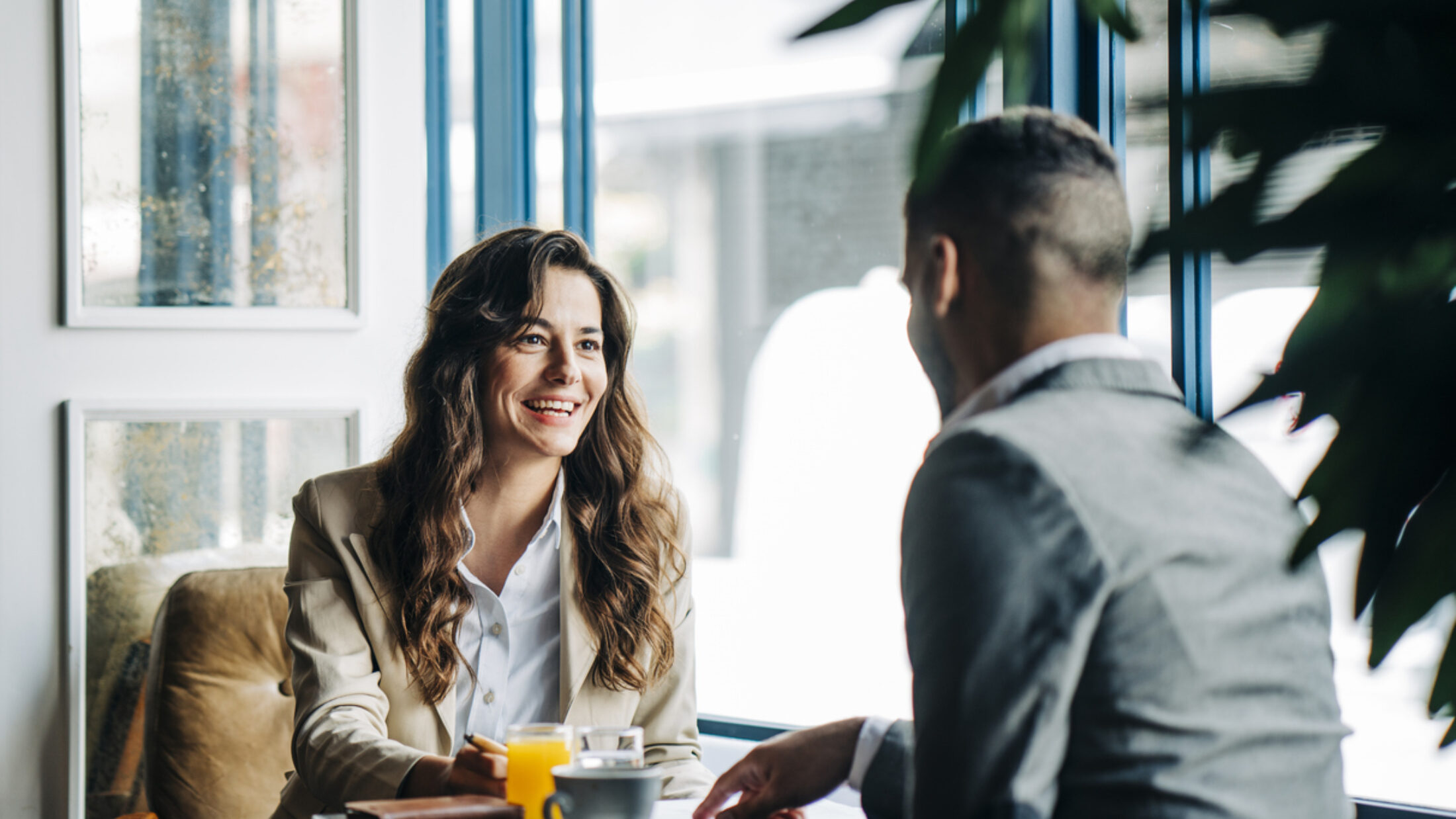 Zwei Personen sitzen in einem Café an einem kleinen Tisch und führen ein Gespräch, während Getränke und Unterlagen vor ihnen liegen.