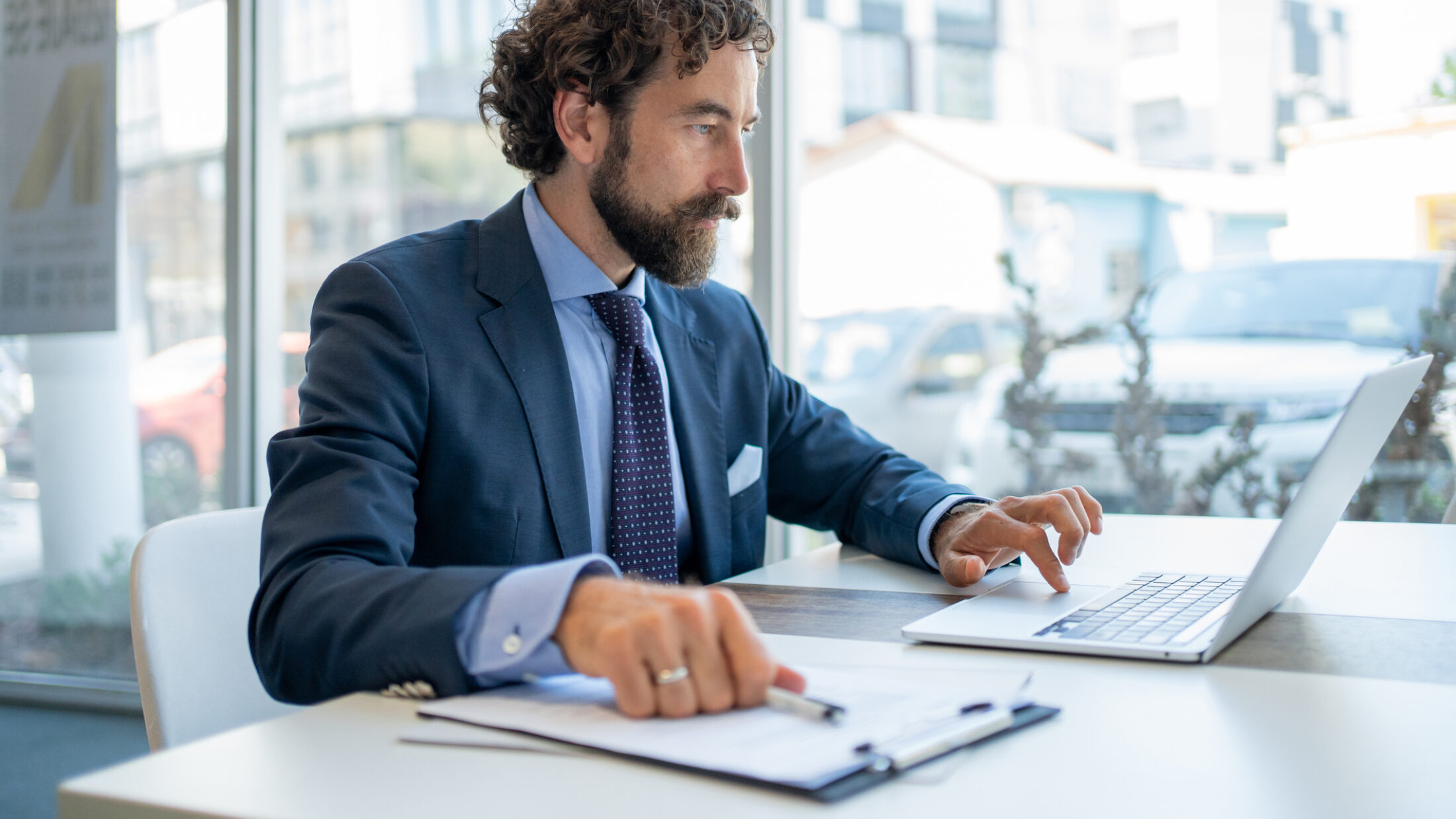 Focused businessman in a suit sitting at a desk, working intently on a laptop while reviewing important documents in a modern office with a city view