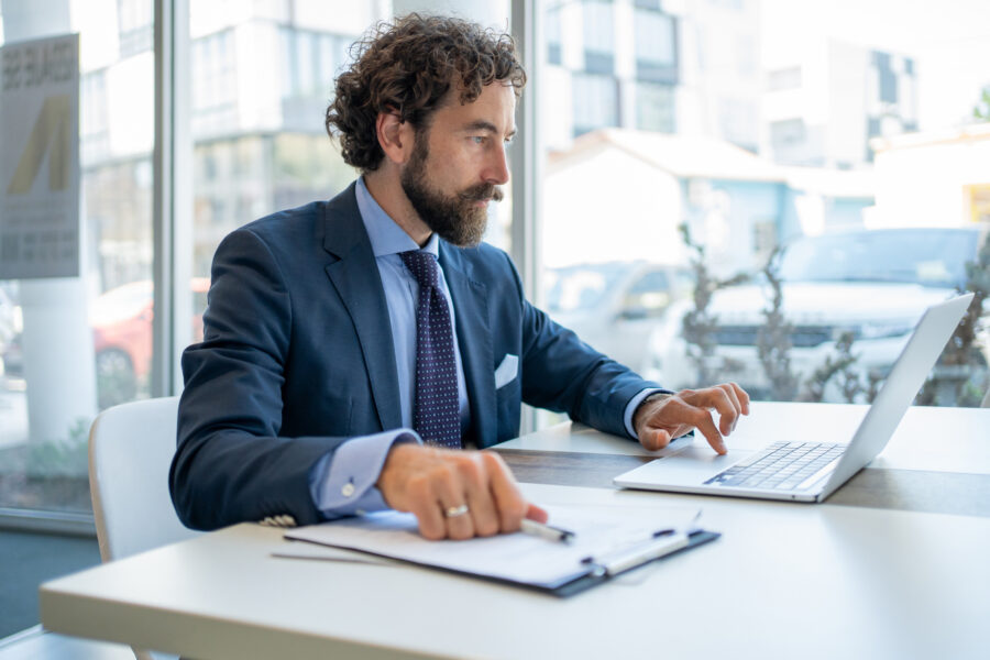 Focused businessman in a suit sitting at a desk, working intently on a laptop while reviewing important documents in a modern office with a city view