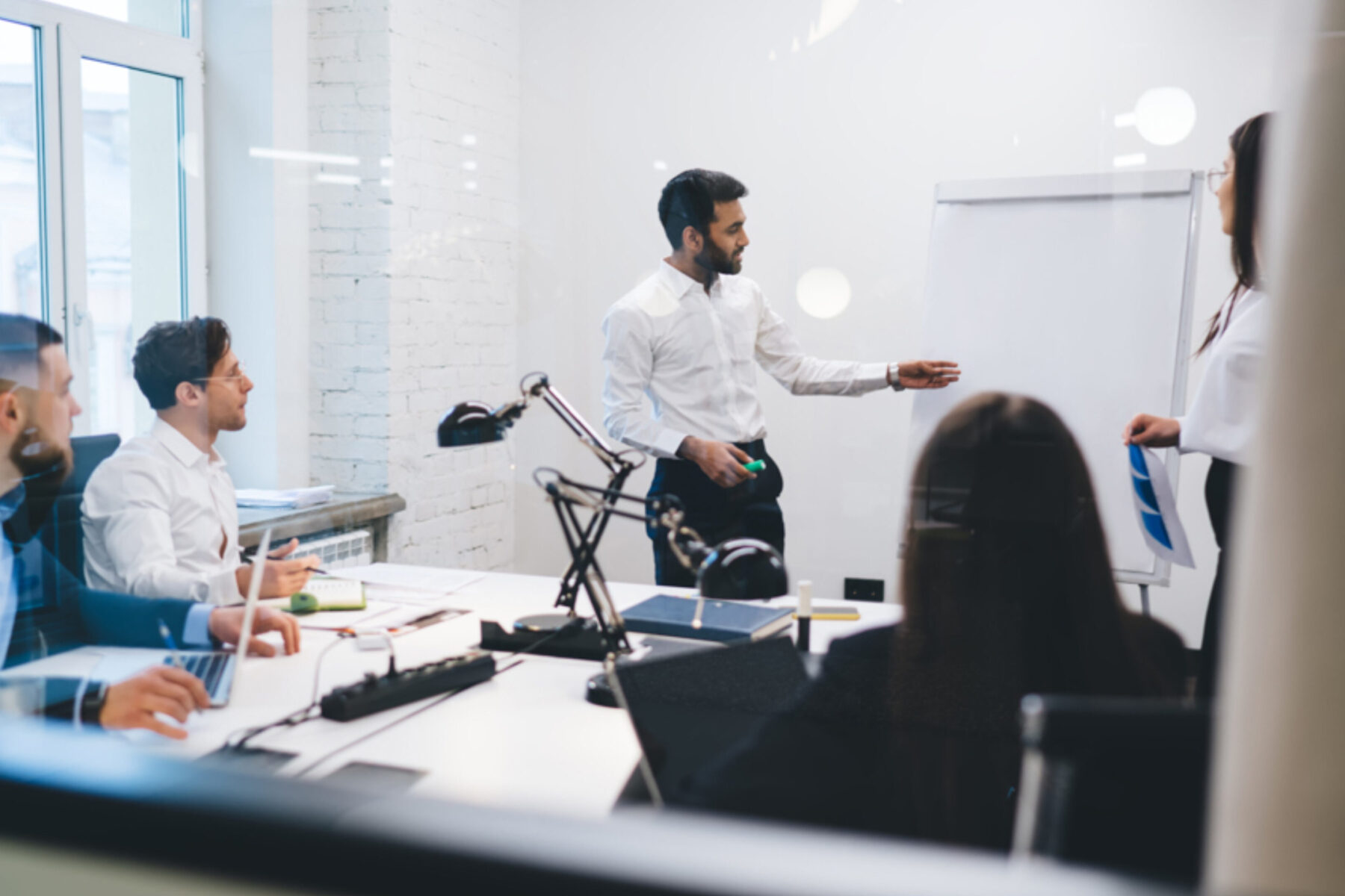 Teammeeting in einem modernen Büro: Ein Mann im weißen Hemd steht an einem Flipchart und präsentiert, während mehrere Kolleginnen und Kollegen am Konferenztisch mit Laptops und Unterlagen sitzen.