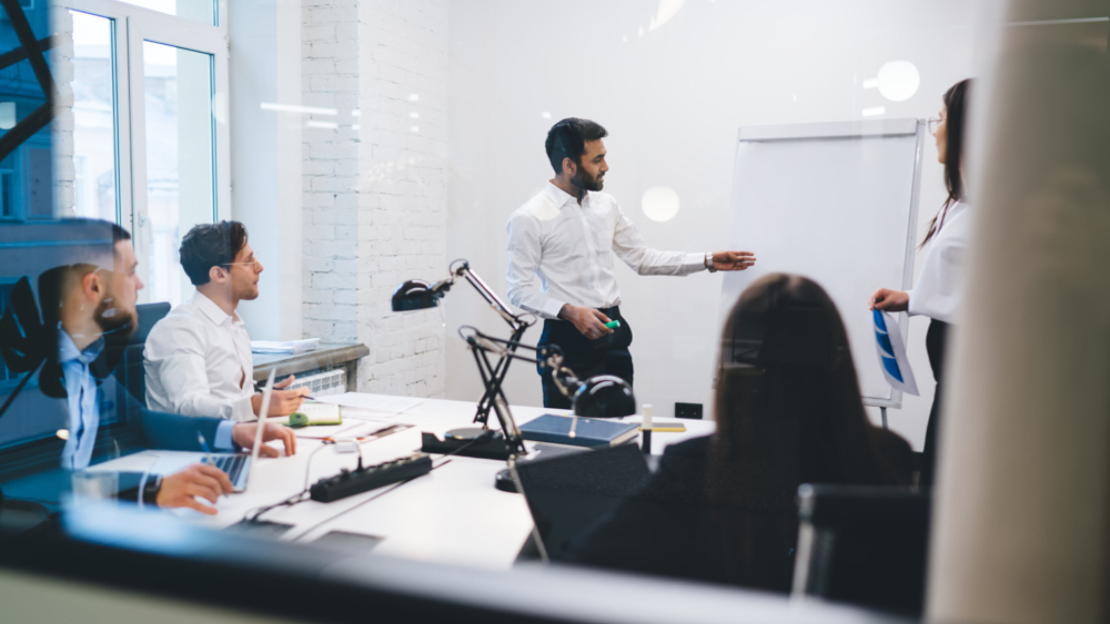 Teammeeting in einem modernen Büro: Ein Mann im weißen Hemd steht an einem Flipchart und präsentiert, während mehrere Kolleginnen und Kollegen am Konferenztisch mit Laptops und Unterlagen sitzen.
